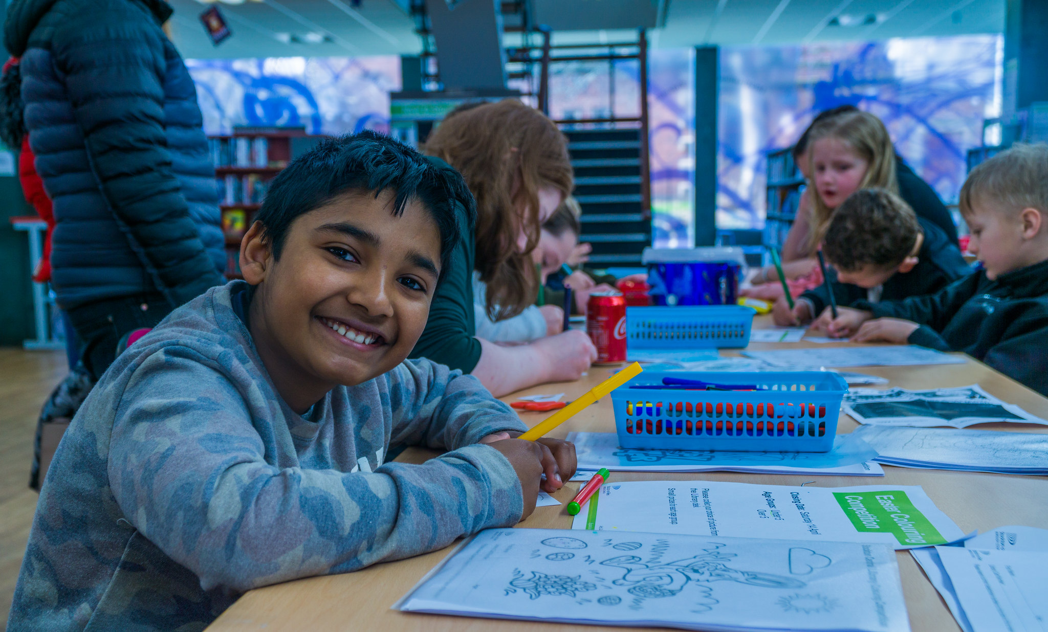 A group of children at a table doing a colouring competition, with a boy in the foreground smiling at the camera.