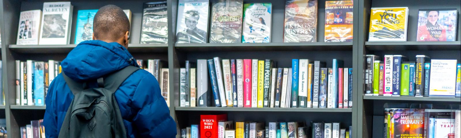 A man with a backpack examines a library shelf full of books.