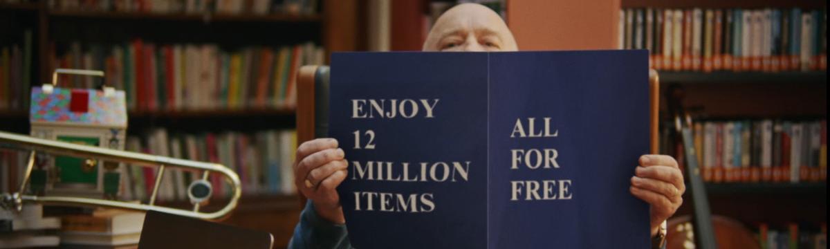 A man sits in the library holding up a magazine, on the cover of the magazine reads "Enjoy 12 million items all for free"