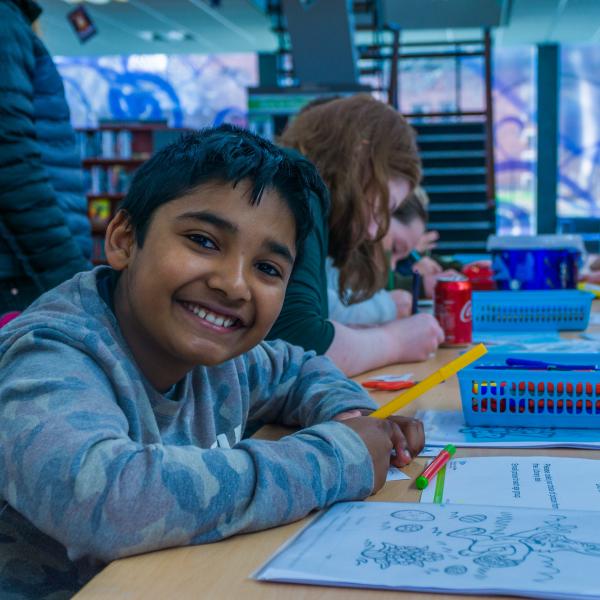 A group of children at a table doing a colouring competition, with a boy in the foreground smiling at the camera.