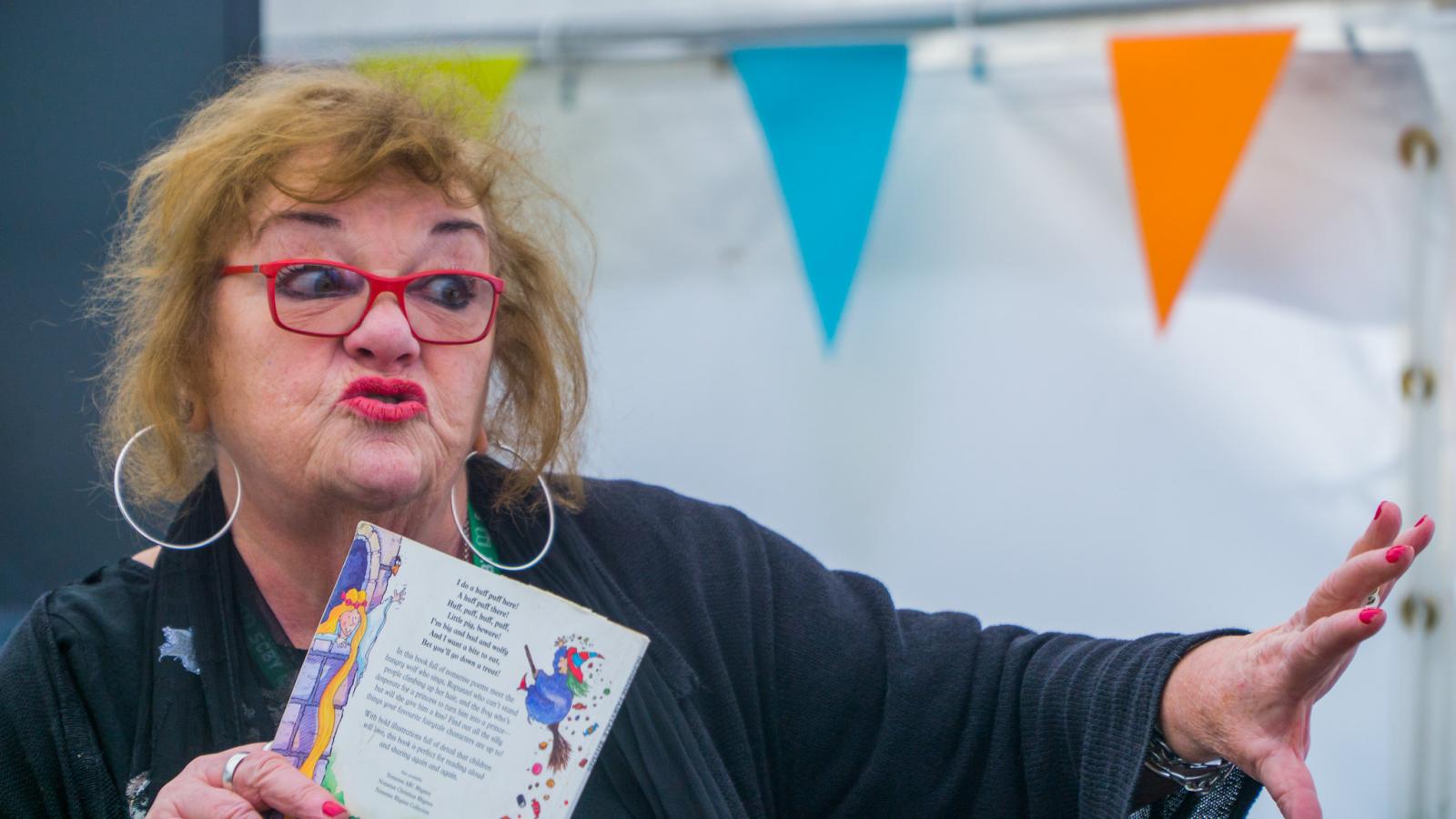 A woman holds a book and makes a dramatic expression and wave of the hand.