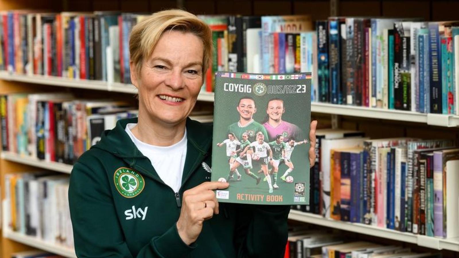 Vera Pauw holds FAI Activity book in front of library shelves
